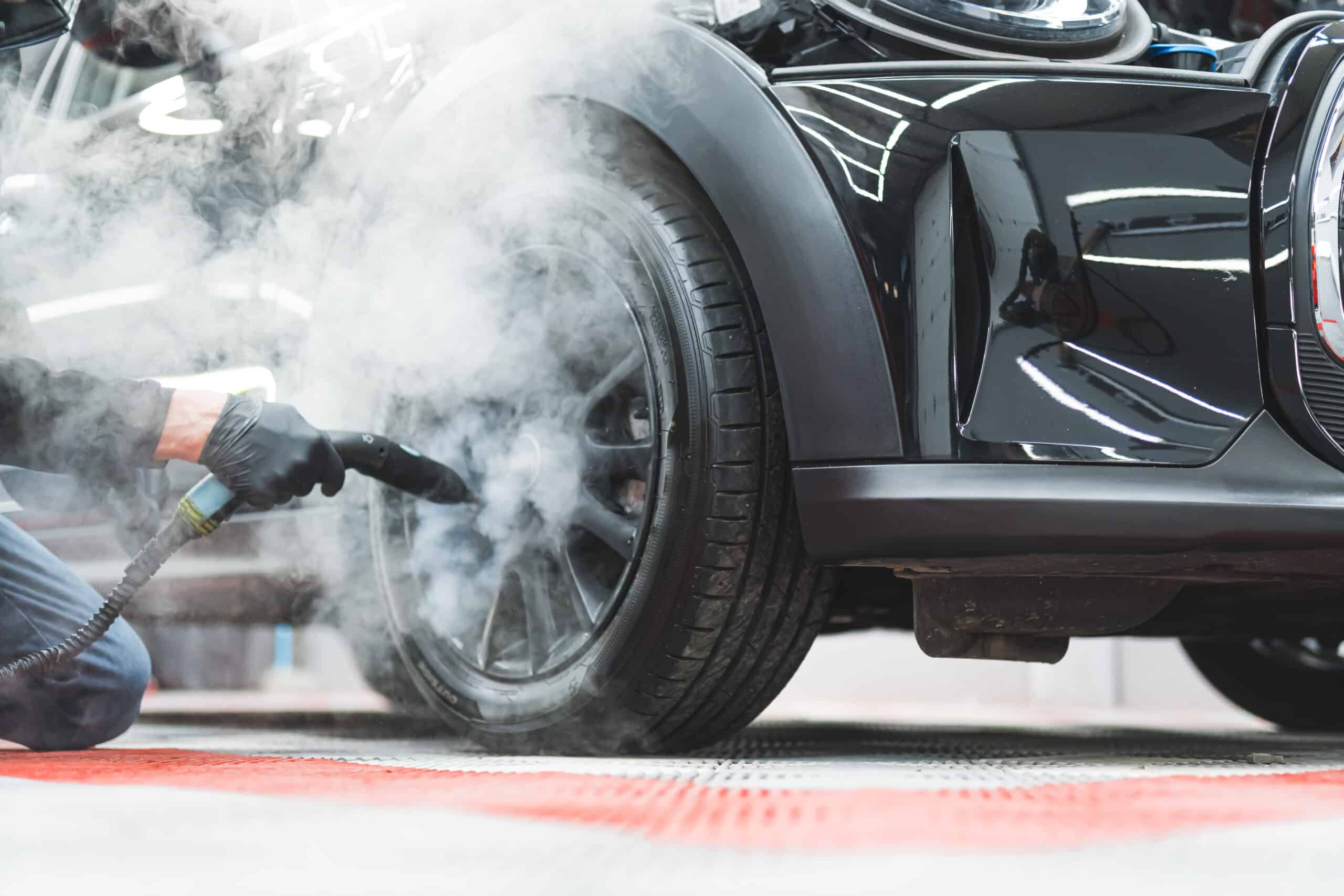Un homme ganté nettoie la jante noire d'une voiture noire à la vapeur, dégageant une épaisse fumée blanche.