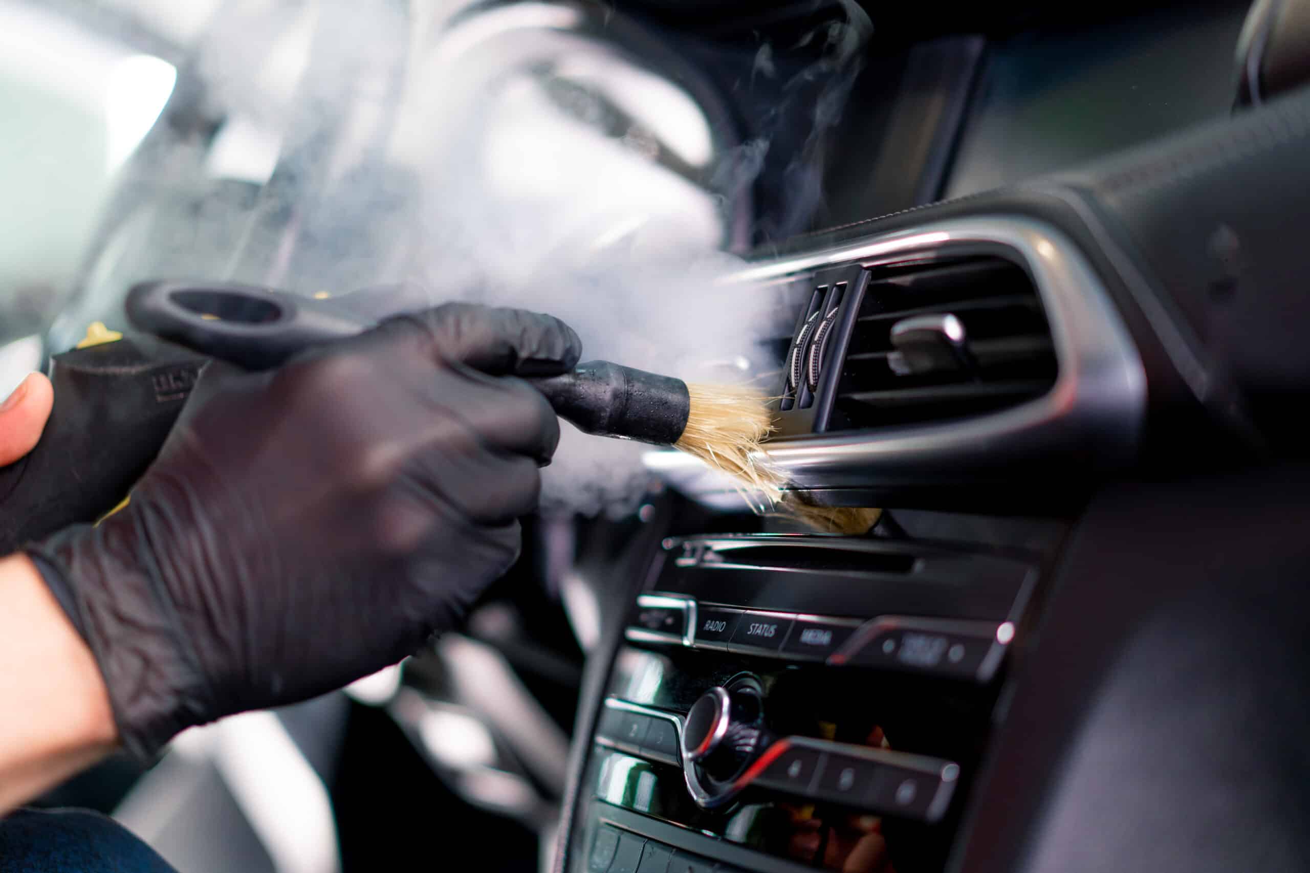Nettoyage vapeur avec brosse d'une bouche d'aération automobile, mains gantées de noir sur tableau de bord près des boutons RADIO, STATUS, MEDIA.