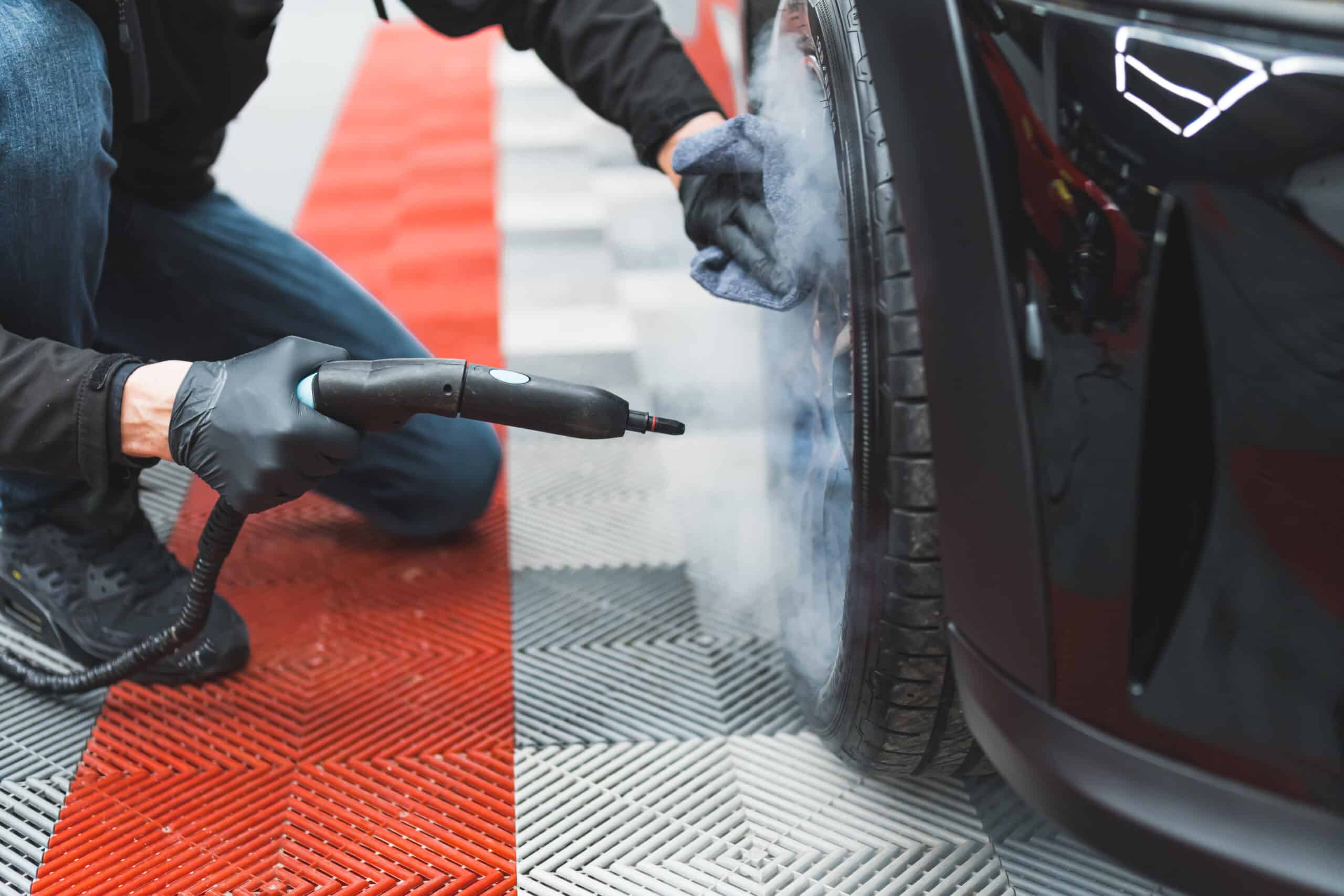 Lavage vapeur jante de voiture par technicien portant des gants noirs sur sol modulaire rouge et gris.