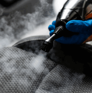 Gros plan sur un nettoyage vapeur d'un siège de voiture en tissu gris foncé, une brosse sort de la vapeur.